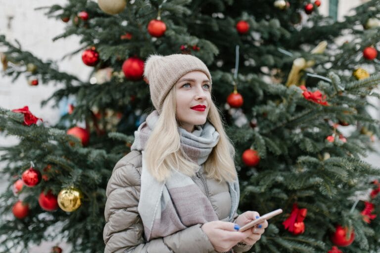 Woman wearing a beige beanie, scarf, and winter jacket standing in front of a decorated Christmas tree, winter outfit 2026