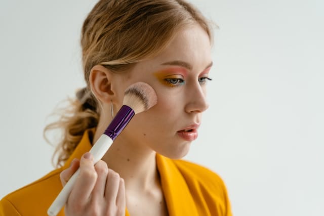 A woman in a yellow blazer using a makeup brush to blend pink cream blush onto her cheekbones for a natural flush